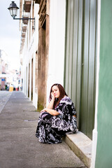 Beautiful, relaxed woman traveling with braids in her hair sitting on a step with her hand on her chin. Old houses in the background.