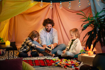 Fototapeta na wymiar A dad is spending time with his two young boys in an indoor blanket fort he is reading out a story to the boys and having a very good time together