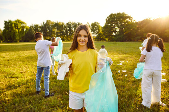 Portrait of a smiling young child girl and children volunteers with gloves and garbage bags cleaning plastic trash from grass in the summer park outdoors. Environmental pollution and ecology concept.