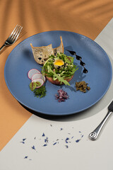 Salmon tartar served on a blue plate with fresh quail egg and fresh greens and bread 
