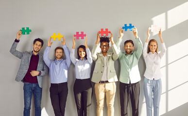 Group of young happy business people holding colorful puzzle parts. Coworkers, colleagues or team of staff standing in a raw together with jigsaw pieces with hands up on a gray wall background. - obrazy, fototapety, plakaty