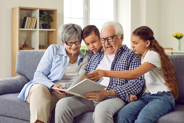 Portrait of a happy grandparents reading a book to their grandchildren or looking through a family photo album sitting on sofa at home. Kids enjoying leisure time with grandma and grandpa on weekend - obrazy, fototapety, plakaty