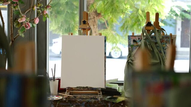 Empty artist canvas on wooden easel and paint brushes near window at bright art studio