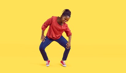 Naklejka na ściany i meble Happy young woman dancing against a vivid vibrant yellow colour studio background. Cheerful beautiful plus size African American dancer girl in comfortable jeans doing lively knee moves to music