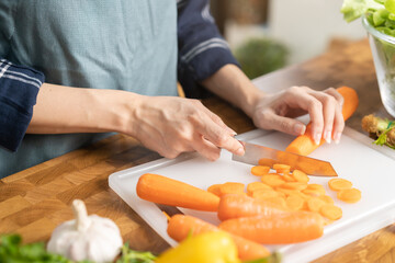 Obraz na płótnie Canvas Asian young woman, girl or housewife hand using knife, cutting carrots on board, on wooden table in kitchen home, preparing ingredient, recipe fresh vegetables for cooking meal. Healthy food people.