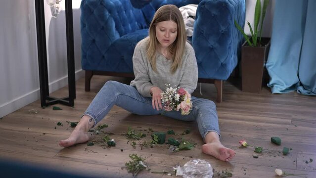 Young angry woman sitting in living room with broken bouquet crying. Wide shot portrait of stressed desperate Caucasian beautiful lady after fight alone at home indoors