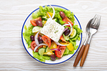 Traditional Greek Salad with Feta Cheese, Tomatoes, Bell Pepper, Cucumbers, Olives, Herbs in white ceramic bowl on White rustic wooden table background from above, Cuisine of Greece