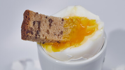 A piece of bread is dipped into an egg with liquid yolk in a white egg cup