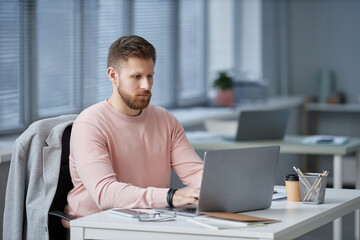 Fototapeta na wymiar Young serious businessman in casualwear looking through online data on laptop screen and typing while networking or preparing report