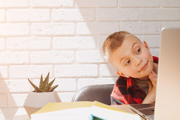 A smiling school-age boy sits at a table with a laptop. Children and gadgets. Front view