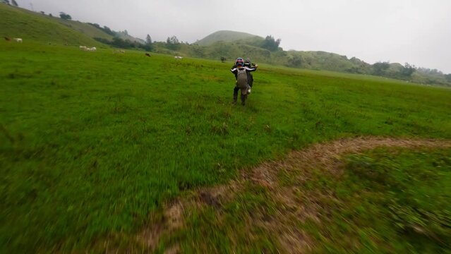 Enduro motorbike biker speed riding extreme stunt tricks wheely on green valley field aerial view. FPV sport drone close shot male racer standing on back wheel motorcycle adrenalin travel adventure 4k