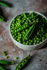 Bowl with fresh sweet peas with two open pods on the top on stone surface.