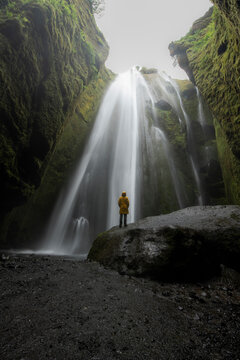 View of a girl in a yellow rain coat standing on a rock below the waterfall in Iceland.