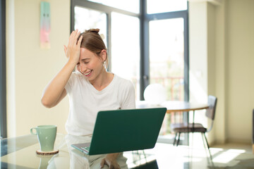 caucasian pretty woman with a laptop laughing and slapping forehead like saying d’oh! I forgot or...