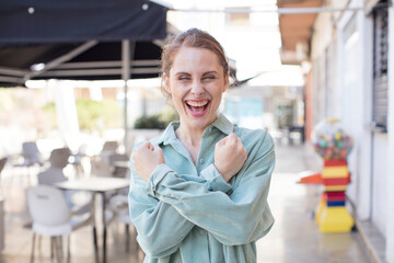 pretty young woman smiling cheerfully and celebrating, with fists clenched and arms crossed, feeling happy and positive