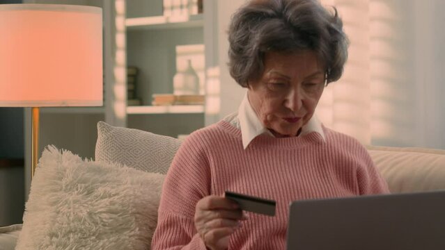Happy Caucasian old woman senior granny elderly female mature lady retired grandmother with laptop and bank credit card paying online shopping with computer buying internet payment celebrate at home