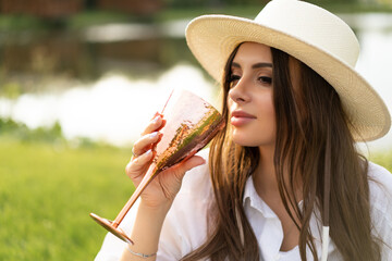beautiful glamorous girl in a white dress and a straw hat on the shore of the lake with a goblet in...