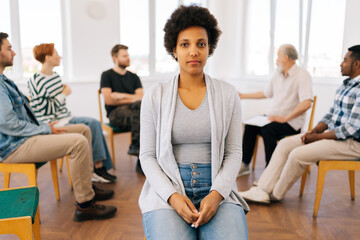 Portrait of pretty African young woman looking at camera with serious expression during group therapy session, multicultural and different ages people sitting in circle on background.