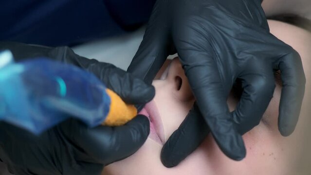 Permanent makeup procedure applying rose red pigment using tattoo machine in beautician salon. Microblading tattoo lips.