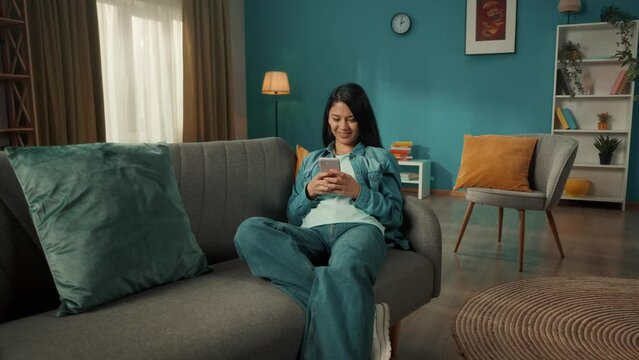 A young Asian woman sits on a sofa with a phone in her hands. A woman is typing a message, chatting, browsing social networks, scrolling through the feed.