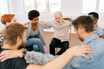 Top view of mature male therapist and group of multi-age patients putting hands on each others shoulders sitting in circle during group therapy session as symbol of support. Concept of mental health.