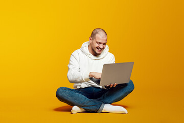 Young guy sitting on floor and using modern notebook, typing on laptop and smiling, isolated on...