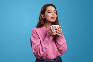Pretty young girl enjoying aroma of coffee with eyes closed, holding white cup of hot beverage, isolated over blue background