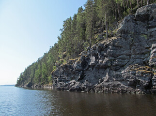 Beautiful summer nature of northern lake. Landscape with rocky coast, green trees, blue sky and reflection in calm water. Ladoga lake, Karelia, Russia.