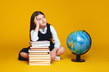 Exhausted young schoolgirl holding hand on head, sitting near books and earth globe while looking away, isolated over yellow background