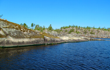 Rocky coast with large stones, trees and reflection in quiet water surface of northern lake. Northern nature and blue sky in summer. Ladoga lake, Karelia, Russia.