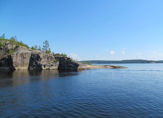 Rocky coast with large stones, trees and reflection in quiet water surface of northern lake. Northern nature and blue sky in summer. Ladoga lake, Karelia, Russia.