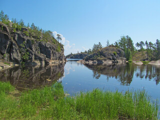 Fototapeta na wymiar Rocky coast with large stones, trees and reflection in quiet water surface of northern lake. Northern nature and blue sky in summer. Ladoga lake, Karelia, Russia.