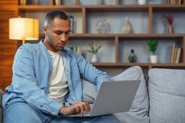 Handsome african man working on laptop computer while sitting on a sofa in stylish cozy living room. Freelancer working fom home. Browsing internet, using social networks