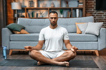 Young man meditating on his living room floor sitting in the lotus position with his eyes closed and an expression of tranquility in a health and fitness concept - 624474885