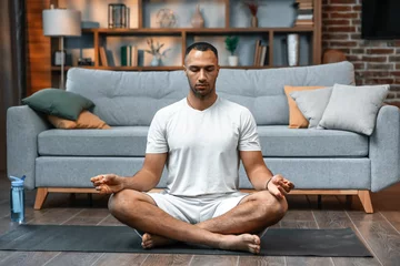 Foto op Canvas Young man meditating on his living room floor sitting in the lotus position with his eyes closed and an expression of tranquility in a health and fitness concept © Ivan