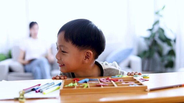 Lifestyle naughty hyperactive boy playing and looking to camera wtihe colorful toy number in front of him, Asian mother reading book in living room, mother and cute son spend time together at home