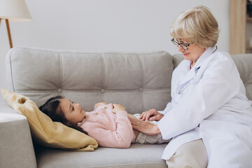 Senior female doctor in white coat palpating belly of little girl lying on couch