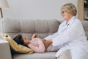 Senior female doctor in white coat palpating belly of little girl lying on couch - obrazy, fototapety, plakaty