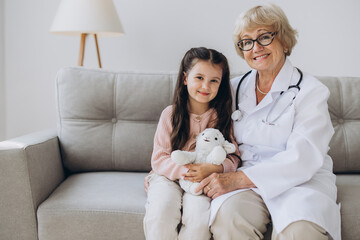 Portrait of senior nurse wearing white coat, embracing shoulders of happy little preschool patient....