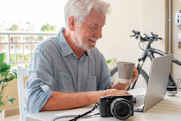 Happy white haired senior man sitting outdoor on terrace holding a coffee cup while typing on...