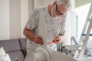 Elderly bearded man works to install a paddle fan on the ceiling at home, experienced senior electrician installs a fan appliance to beat the heat