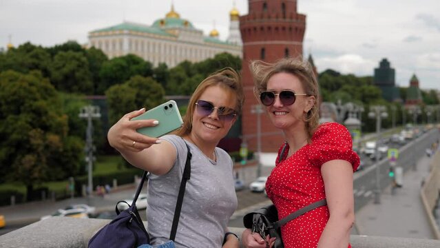 Two smiling young women in sunglasses take selfies standing on a bridge against the background of a river and a busy road. Two tourists take pictures of the sights of the city.