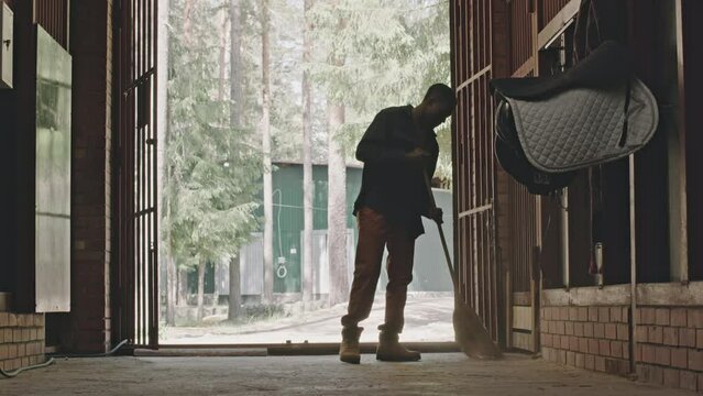 Full shot of young black man sweeping floor in horse stable while doing cleanup on farmFull shot of young black man sweeping floor in horse stable while doing cleanup on farmFull shot of young black m