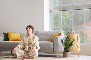 Pretty young woman lying meditating near grey sofa in light living room - Powered by Adobe