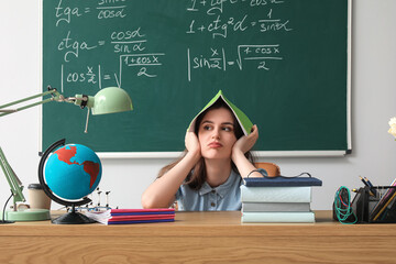 Tired female teacher with copybook sitting at table in classroom