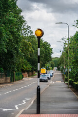 Alerting yellow traffic light at crosswalk on English road, against cloudy sky and green trees. Flashing light signals presence of crosswalk. Linear and aerial perspective. Vertical crop.