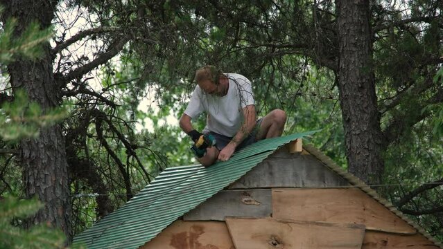 Close up of man securing green sheets of metal onto roof of tree house build from scarp material around two trees.