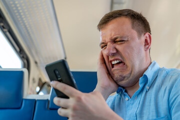 Disgusted and overwhelmed man stares at screen of smartphone with confuse, bad joke or inappropriate content. Sad millennial guy looks at phone screen in surprise, sits in commuter train. - obrazy, fototapety, plakaty