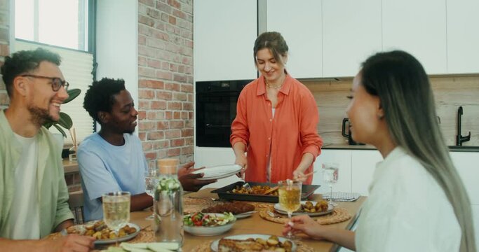 A woman puts fried potatoes and meat on the plates of her friends during a joint dinner at home