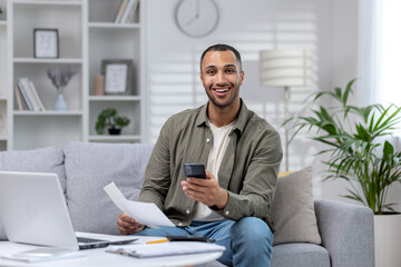 Portrait of a young African-American businessman who works remotely from home. He is sitting on the sofa in front of the laptop, holding a phone and documents in his hands. Smiling at the camera